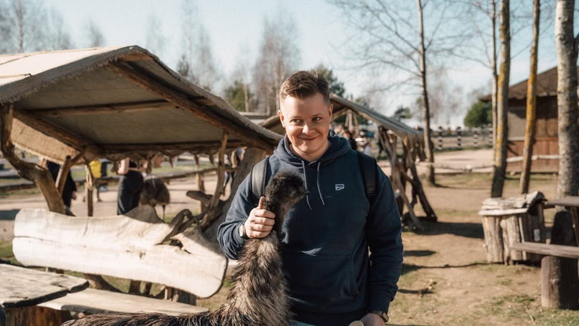 A cheerful young man interacting with an emu at a rustic outdoor farm setting, highlighting rural life.